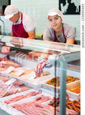 Female seller leans over to refrigerator window and takes out some raw barbecue sausages for buyer 133093993