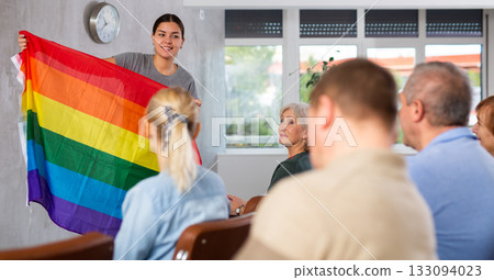 Portrait of young female teacher conducting lesson for adult students, talking about LGBT community and showing rainbow flag 133094023