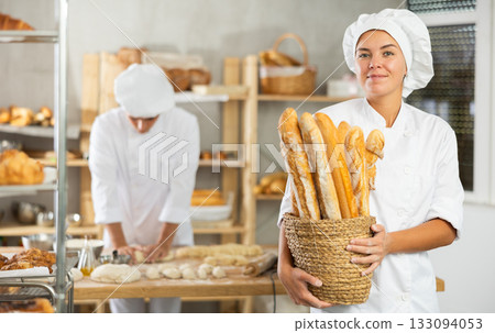 Young female baker holding baguettes in round wicker basket 133094053