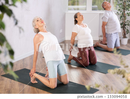 Elderly women together with a young coach doing stretching and doing yoga on mats in gym Elderly women together with a young coach doing stretching and doing yoga on mats in gym 133094058