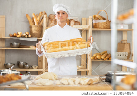 Young male baker holding baguettes in wicker basket Young male baker holding baguettes in wicker basket 133094094