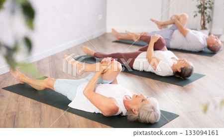 At yoga class elderly woman is lying on back, bent one leg at knee and raised it to chest 133094161