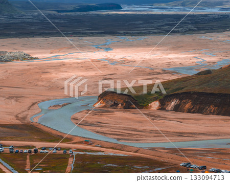 Sweeping aerial view of a winding river through a sandy plain, with a rocky hill, parked vehicles, dirt tracks, and distant hills in Iceland. 133094713