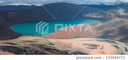 Turquoise crater lake surrounded by dark volcanic terrain, sandy delta, braided rivers, rugged hills, and distant mountains in Iceland. Turquoise crater lake surrounded by dark volcanic terrain, sandy delta, braided rivers, rugged hills, and distant mountains in Iceland. 133094715