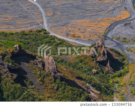 Rugged Icelandic terrain with jagged rocks, lush green vegetation, a winding gravel road, and barren ground with patches of golden moss or grass. Rugged Icelandic terrain with jagged rocks, lush green vegetation, a winding gravel road, and barren ground with patches of golden moss or grass. 133094716