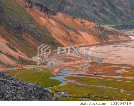 Vibrant orange and brown rhyolite hills with green mossy plains, winding streams, and a road with parked cars in Landmannalaugar, Iceland. 133094719