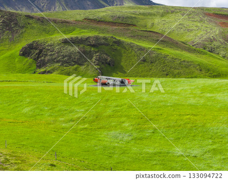 An abandoned Abandoned U.S. Navy Douglas C 117D aircraft fuselage with orange accents rests on vibrant green terrain, surrounded by rolling hills and rocky outcrops in Iceland. 133094722