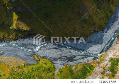 Aerial perspective of a winding river with dark volcanic gravel cutting through a green canyon with steep cliffs and vibrant vegetation in Iceland. 133094726