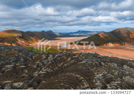 Aerial view of Iceland's Landmannalaugar region with vibrant rhyolite mountains, dark lava fields, a winding river, and a cloudy sky in the highlands. 133094727