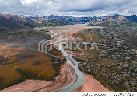 Aerial view of a winding river in Iceland's colorful Landmannalaugar region, with volcanic rock formations, snow capped mountains, and a partly cloudy sky. 133094729