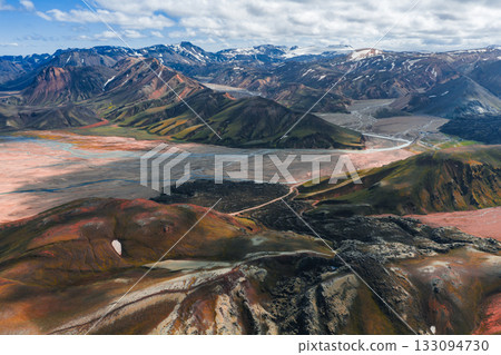 Aerial view of vibrant rhyolite mountains in Landmannalaugar, Iceland, featuring red, orange, green, and brown hues, snow patches, and a winding river. 133094730