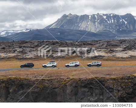 Four off road vehicles travel along a dirt path in Iceland, with a snow patched volcanic mountain and rocky terrain under an overcast sky. Four off road vehicles travel along a dirt path in Iceland, with a snow patched volcanic mountain and rocky terrain under an overcast sky. 133094735
