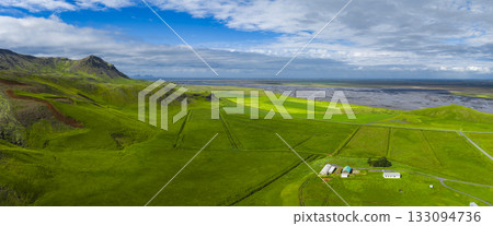 Vibrant green fields with white and green roofed farm buildings, a rugged mountain on the left, and a flat coastal plain under a partly cloudy sky in Iceland. Vibrant green fields with white and green roofed farm buildings, a rugged mountain on the left, and a flat coastal plain under a partly cloudy sky in Iceland. 133094736