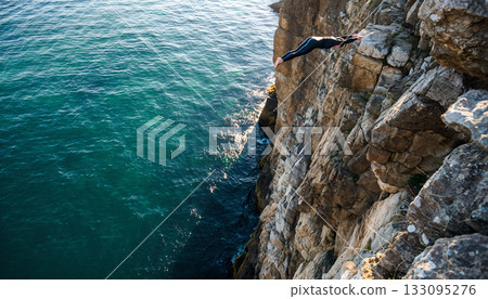 Daredevil individual executing a breathtaking cliff dive toward the turquoise ocean showcasing 133095276