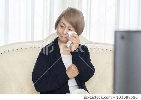 一位老婦人在客廳一邊看悲傷的電視節目一邊哭泣 一位老婦人在客廳一邊看悲傷的電視節目一邊哭泣 133098586