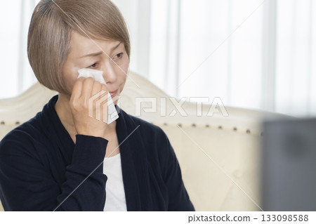 一位老婦人在客廳一邊看悲傷的電視節目一邊哭泣 133098588