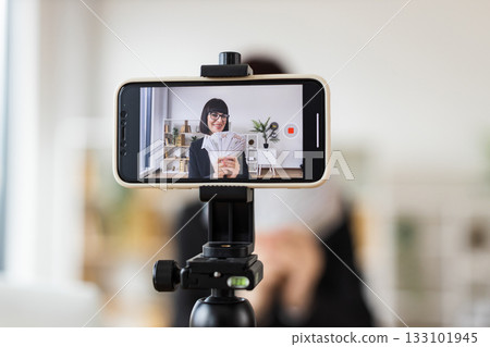 Caucasian woman mid 30s recording video blog about investments, financial literacy, income, holding cash, sitting at desk in modern office environment with smartphone on tripod for content creation 133101945