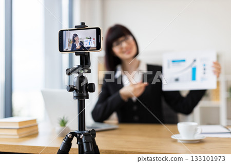 Caucasian woman recording business blog using smartphone on tripod in office environment. Discussing chart data in video. Professional creating online content. Relates to business, content creation. Caucasian woman recording business blog using smartphone on tripod in office environment. Discussing chart data in video. Professional creating online content. Relates to business, content creation. 133101973