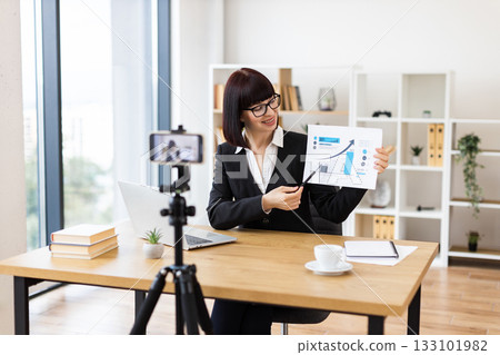 Caucasian woman sitting at table recording blog video about business performance statistics and growth analytics. Caucasian woman sitting at table recording blog video about business performance statistics and growth analytics. 133101982