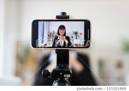 Caucasian adult woman sitting at desk, recording business blog with smartphone camera. Image shows professional woman in modern office environment creating content about business topics. 133101989