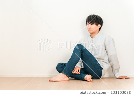 一位身穿牛仔褲的年輕亞裔男子坐在白牆的客廳裡 一位身穿牛仔褲的年輕亞裔男子坐在白牆的客廳裡 133103088