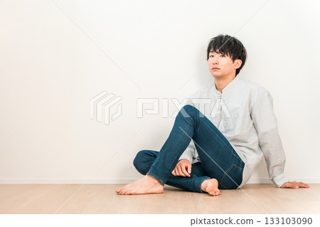 一位身穿牛仔褲的年輕亞裔男子坐在白牆的客廳裡 一位身穿牛仔褲的年輕亞裔男子坐在白牆的客廳裡 133103090