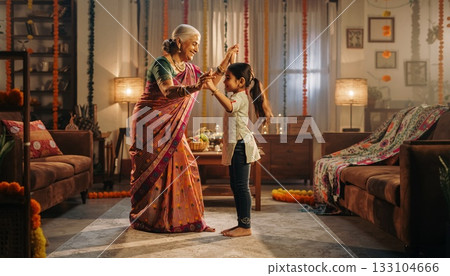 Grandmother in Traditional Sari Dancing with Young Granddaughter During Festive Cultural Celebration at Home 133104666