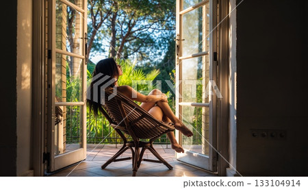 Contemplative Woman Relaxing in Wicker Chair by Open French Doors with Tropical Garden View 133104914