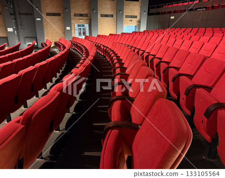 Rows of red theater seats with warm lighting. Architectural rhythm, symmetry, and cultural anticipation reflected in calm interior geometry. Rows of red theater seats with warm lighting. Architectural rhythm, symmetry, and cultural anticipation reflected in calm interior geometry. 133105564