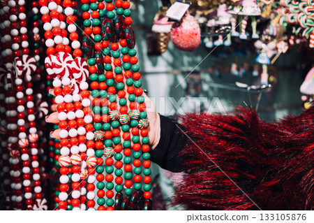 Close-up of a woman hand touching colorful Christmas candy garlands at a festive market stall. Peppermint decor, edible-look ornaments, sweet-themed holidays, dessert-inspired design trends Close-up of a woman hand touching colorful Christmas candy garlands at a festive market stall. Peppermint decor, edible-look ornaments, sweet-themed holidays, dessert-inspired design trends 133105876