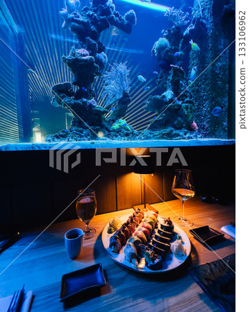 Large sushi platter with various colorful rolls served on wooden table in modern restaurant with illuminated aquarium and glasses of wine and beer, creating luxurious dining experience. Upscale dining 133106962