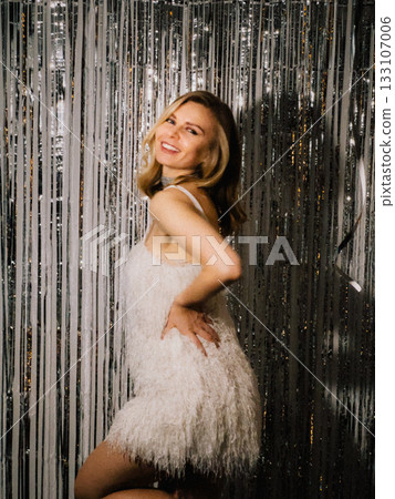 Smiling woman wearing white dress, posing gracefully near shimmering silver curtain, radiating joy and excitement during lively New Years Eve party celebration indoors. Elegant woman in white dress Smiling woman wearing white dress, posing gracefully near shimmering silver curtain, radiating joy and excitement during lively New Years Eve party celebration indoors. Elegant woman in white dress 133107006