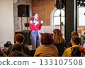 Woman speaks on stage in red off shoulder top to audience during indoor event 133107508
