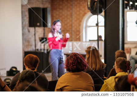 Woman speaks on stage in red off shoulder top to audience during indoor event Woman speaks on stage in red off shoulder top to audience during indoor event 133107508