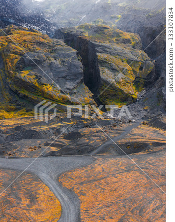 A dramatic Icelandic canyon with steep moss covered cliffs, a gravel path cutting through orange tinged terrain, and dark volcanic rock formations. 133107834