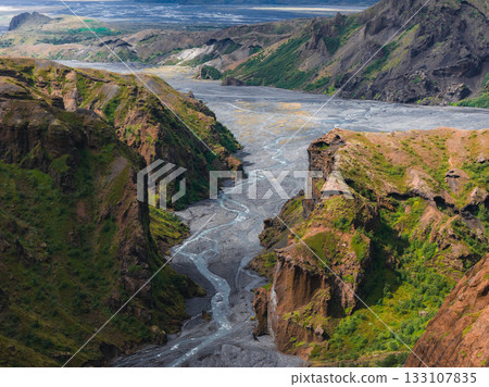Steep cliffs with green vegetation surround a glacial river in Mulagljufur Canyon, Iceland. Plains and mountains stretch across the background. 133107835