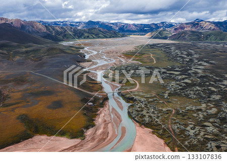 Aerial view of a braided river winding through colorful rhyolite mountains, mossy vegetation, and volcanic rock in Iceland's Landmannalaugar region. 133107836