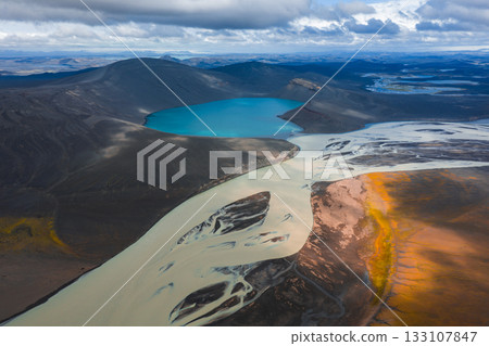A turquoise lake with braided sediment laden rivers flows through dark volcanic terrain, contrasted by orange and yellow patches under dramatic clouds. A turquoise lake with braided sediment laden rivers flows through dark volcanic terrain, contrasted by orange and yellow patches under dramatic clouds. 133107847