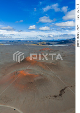 Barren volcanic terrain in Iceland with reddish orange craters, distant mountains, and a small vehicle on a dirt road under a blue sky. Barren volcanic terrain in Iceland with reddish orange craters, distant mountains, and a small vehicle on a dirt road under a blue sky. 133107891