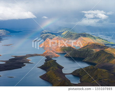 Aerial view of Iceland's colorful rhyolite mountains, interconnected lakes, and a vibrant rainbow under dramatic lighting with scattered clouds. Aerial view of Iceland's colorful rhyolite mountains, interconnected lakes, and a vibrant rainbow under dramatic lighting with scattered clouds. 133107893