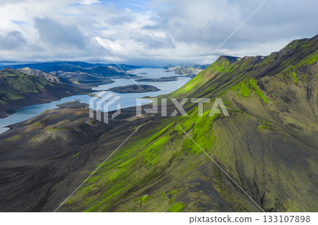 Aerial view of green moss covered hills and dark volcanic terrain in Iceland, with elongated lakes and steep mountain ridges under a partly cloudy sky. Aerial view of green moss covered hills and dark volcanic terrain in Iceland, with elongated lakes and steep mountain ridges under a partly cloudy sky. 133107898