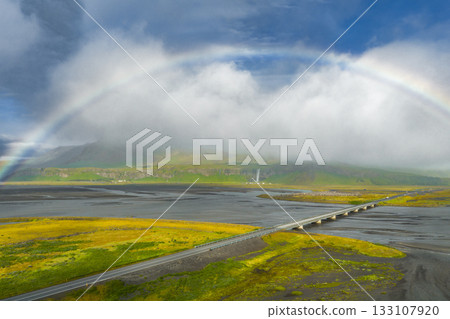 Aerial view of a bridge crossing a wide river in Iceland, surrounded by mossy terrain. A full rainbow arcs above, with clouds over distant cliffs. Aerial view of a bridge crossing a wide river in Iceland, surrounded by mossy terrain. A full rainbow arcs above, with clouds over distant cliffs. 133107920
