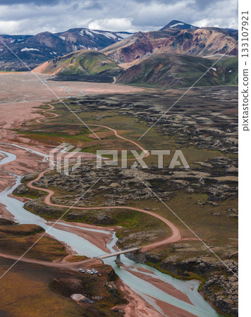 Aerial view of glacial rivers winding through rocky terrain, a small bridge, a dirt road, and vibrant multicolored hills with snow capped peaks in Iceland. Aerial view of glacial rivers winding through rocky terrain, a small bridge, a dirt road, and vibrant multicolored hills with snow capped peaks in Iceland. 133107921