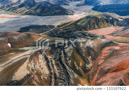 Aerial view of Iceland's Landmannalaugar region with colorful rhyolite mountains, snow patches, volcanic rocks, and a winding dirt road leading to a plain. 133107922