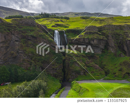 A waterfall flows down a rocky cliff surrounded by green hills under a cloudy sky. A stream winds through the grassy terrain near a visible pathway. A waterfall flows down a rocky cliff surrounded by green hills under a cloudy sky. A stream winds through the grassy terrain near a visible pathway. 133107925