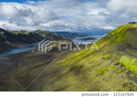 Aerial view of Iceland's moss covered hill, dark volcanic terrain, serene lakes, and jagged mountains under a partly cloudy sky. 133107926
