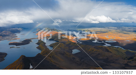 Aerial view of Iceland's highlands featuring multicolored rhyolite mountains, scattered lakes, winding rivers, and a faint rainbow across the scene. 133107937