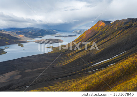 Aerial view of a serene Icelandic lake with irregular islands, volcanic terrain in yellow and orange hues, a faint rainbow, and dramatic clouds. Aerial view of a serene Icelandic lake with irregular islands, volcanic terrain in yellow and orange hues, a faint rainbow, and dramatic clouds. 133107940