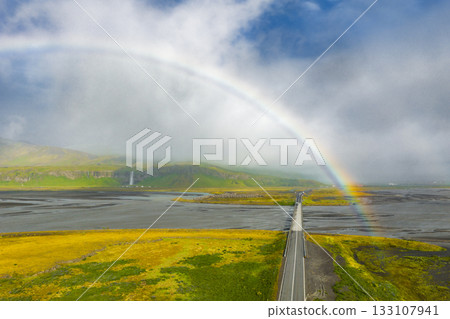 A long road stretches through Iceland's landscape, crossing a gray riverbed. A rainbow arches above, with waterfalls cascading from distant cliffs. A long road stretches through Iceland's landscape, crossing a gray riverbed. A rainbow arches above, with waterfalls cascading from distant cliffs. 133107941