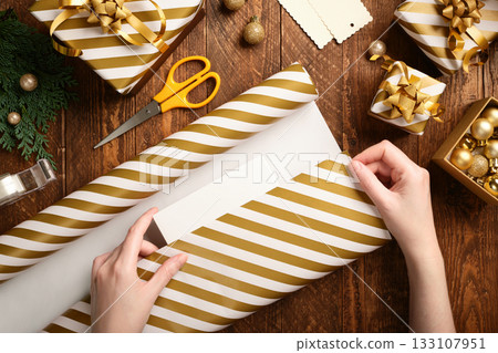 Gift wrapping. Hands wrap a box in striped gold and white paper on a brown wooden table. Gift wrapping. Hands wrap a box in striped gold and white paper on a brown wooden table. 133107951